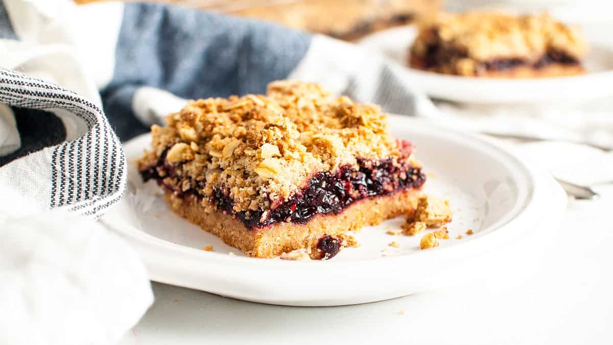 A slice of berry crumble bar with a crumbly oat topping sits on a white plate, surrounded by a striped towel and blurred background elements.