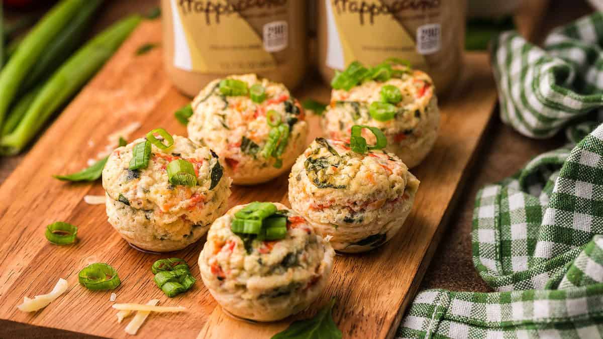 Five vegetable and cheese egg muffins garnished with chopped green onions are arranged on a wooden board, with green onions, shredded cheese, and cloth nearby.