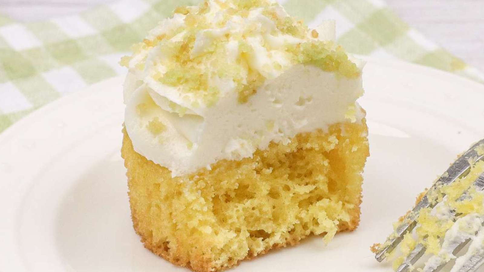 A partially eaten yellow cupcake with white frosting sits on a white plate next to a fork with crumbs.