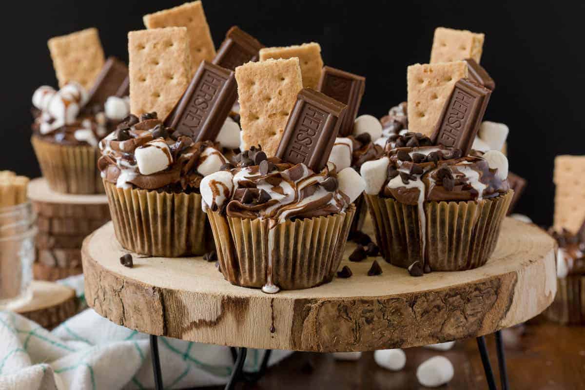 Chocolate cupcakes topped with marshmallows, chocolate bars, graham crackers, and chocolate chips, arranged on a wooden stand.