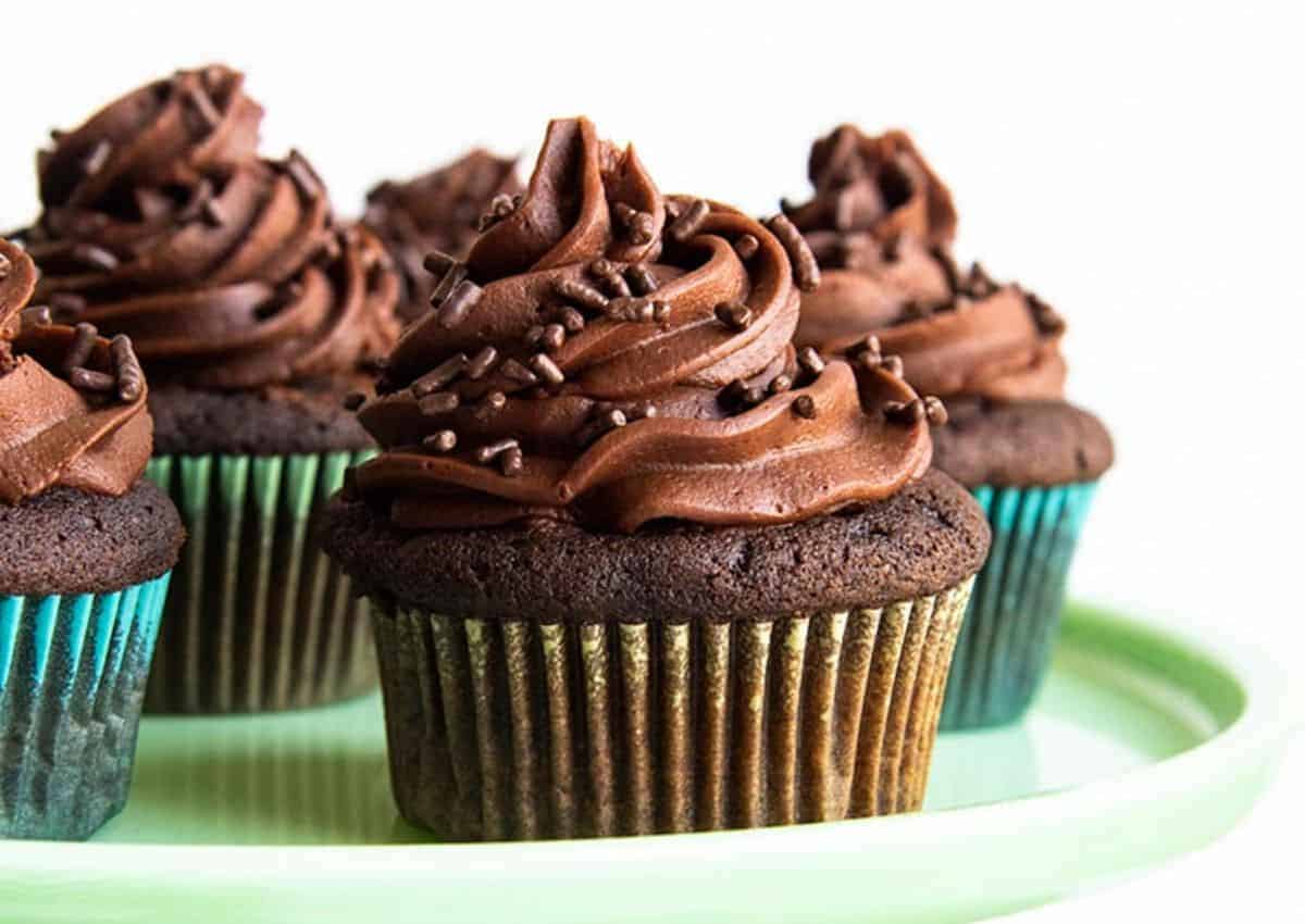 Four chocolate cupcakes with chocolate frosting and chocolate sprinkles are arranged on a light green plate.