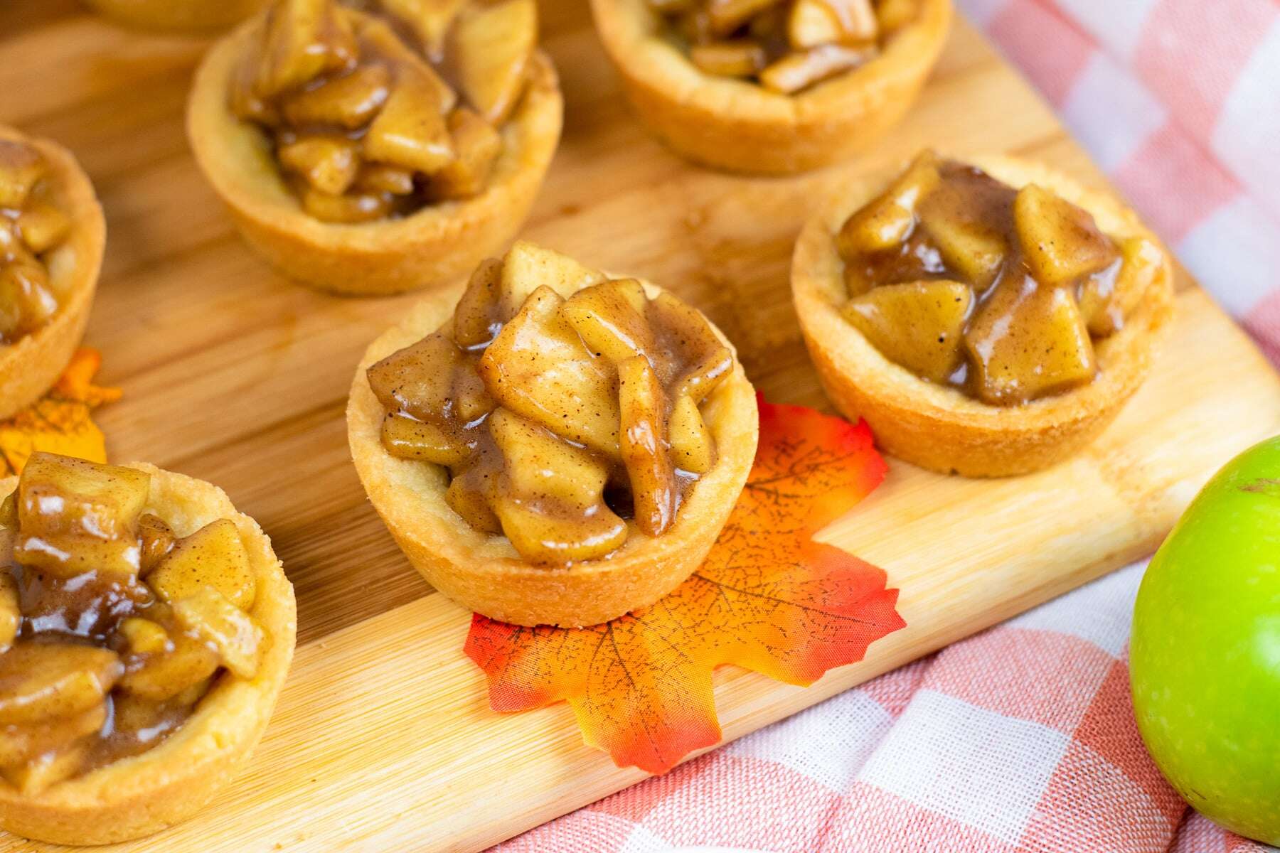 Mini apple pies with spiced apple filling on a wooden board, next to a green apple and a checkered cloth.