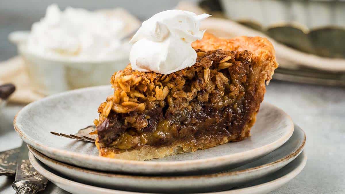 A slice of pie with a crumbly oat topping and a dollop of whipped cream sits on a plate, stacked on another plate with a fork beside it.