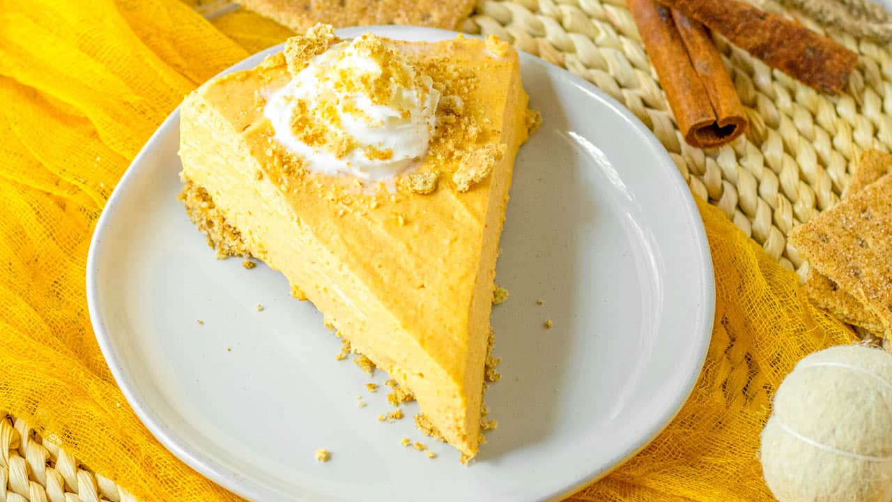 A slice of pumpkin pie with a crumb crust, topped with whipped cream and crumbs, on a white plate with cinnamon sticks and a yellow cloth nearby.