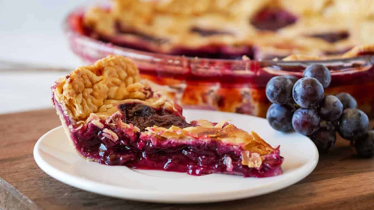 A slice of grape pie sits on a white plate, with a bunch of grapes and the remaining pie in the background.