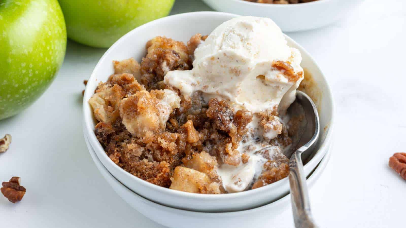 A bowl of apple crisp topped with a scoop of vanilla ice cream, with a spoon inside. Green apples and pecans are visible in the background.