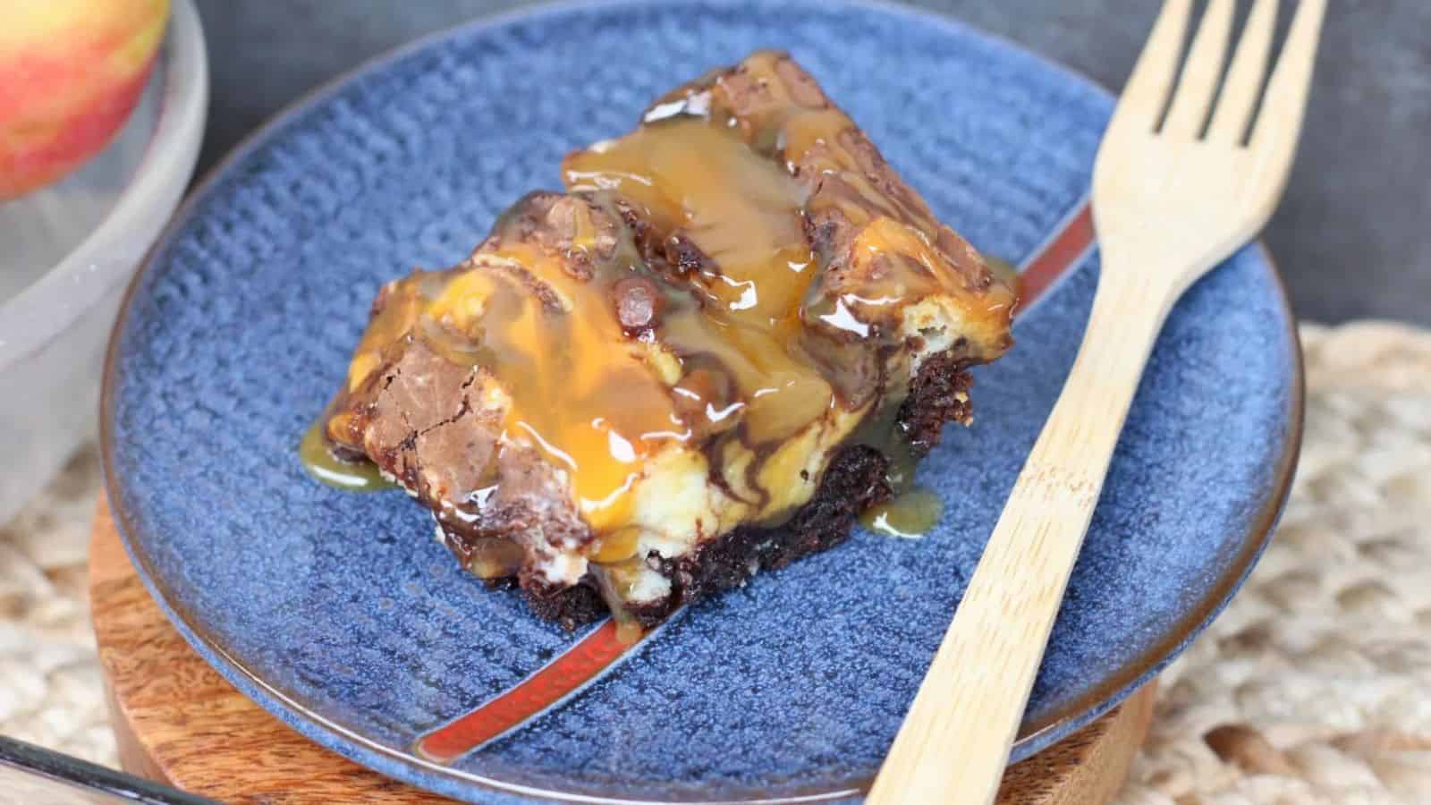 A piece of chocolate brownie with caramel drizzle on a blue plate, next to a wooden fork.
