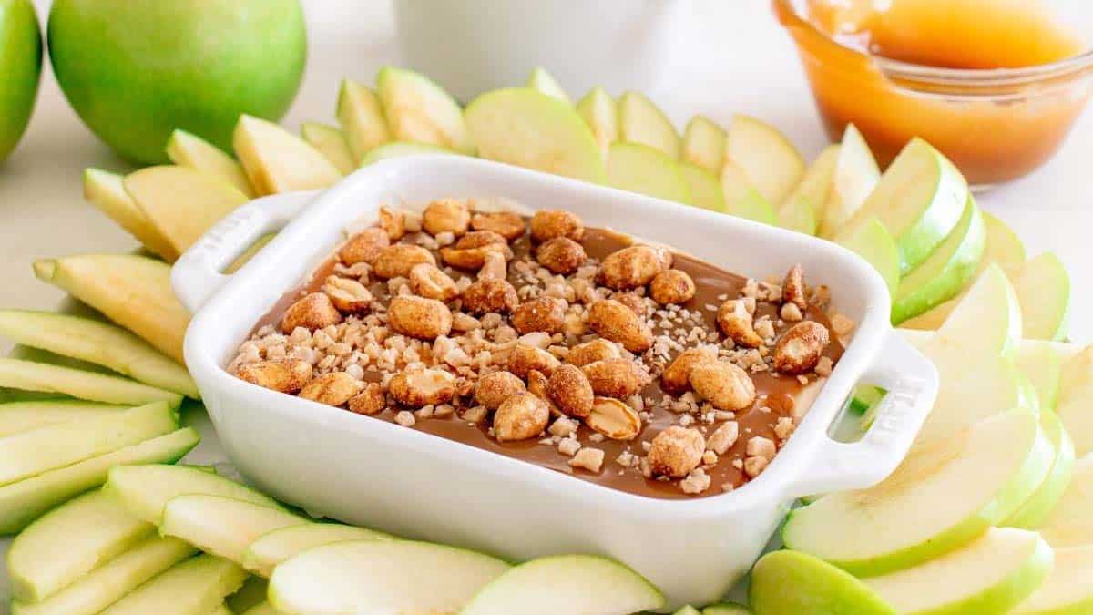 Rectangular white dish filled with caramel dip topped with chopped nuts, surrounded by sliced green apples arranged in a circular pattern.