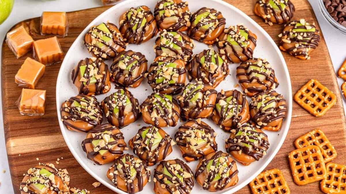 A plate of caramel and chocolate-drizzled pretzel bites topped with chopped nuts, surrounded by caramel squares, chocolate chips, and pretzels on a wooden board.