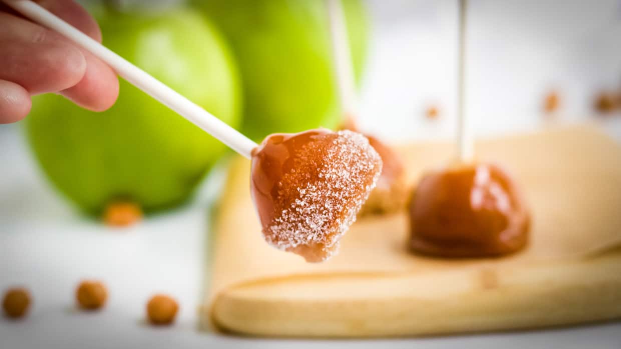 A hand holds a caramel apple pop coated with sugar; more caramel apple pops and green apples are visible in the background.