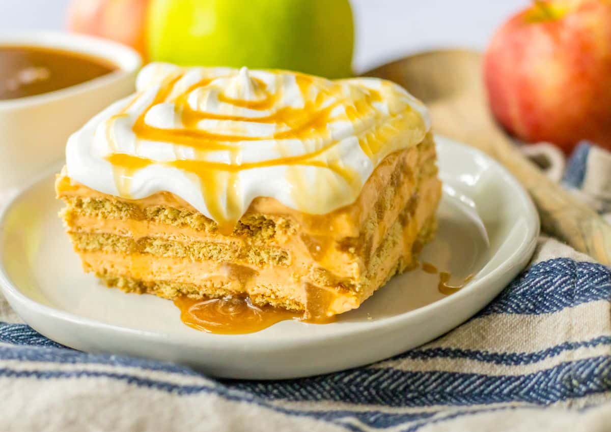 A slice of layered dessert with whipped cream and caramel sauce on top, served on a white plate with apples and caramel sauce in the background.