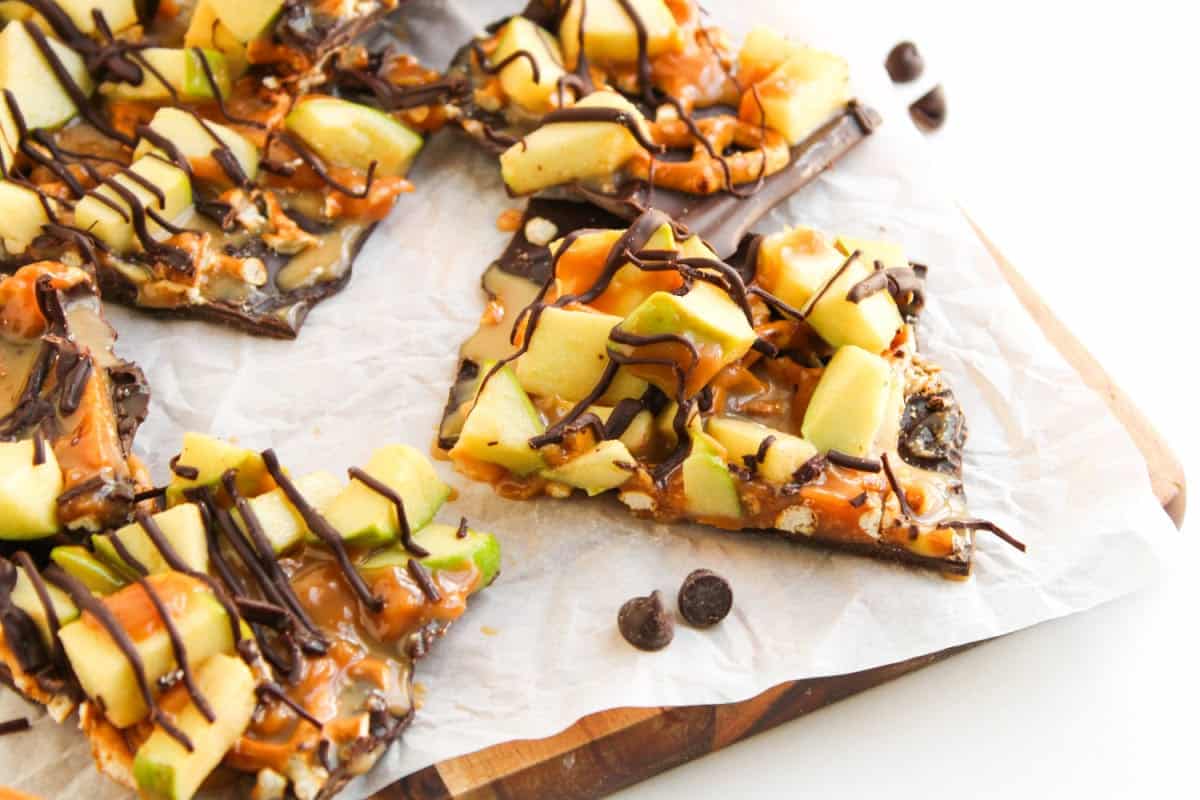Sliced dessert bark topped with chopped green apples, caramel drizzle, and chocolate, served on parchment paper with a few chocolate chips nearby.