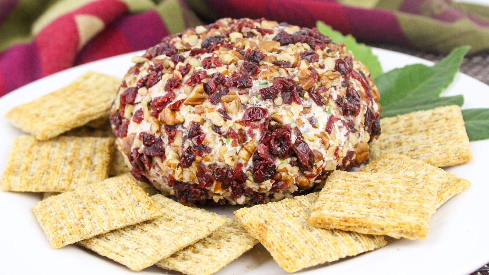 A round cheese ball coated in chopped nuts and dried cranberries is surrounded by rectangular crackers on a white plate.