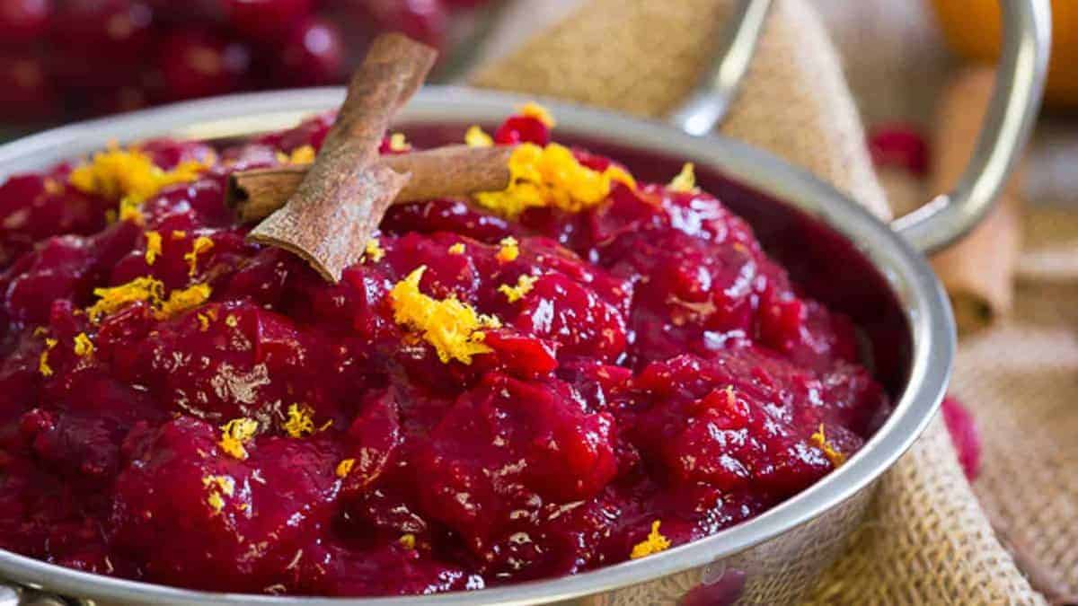 A metal bowl filled with cranberry sauce, garnished with orange zest and two cinnamon sticks, placed on a burlap cloth.