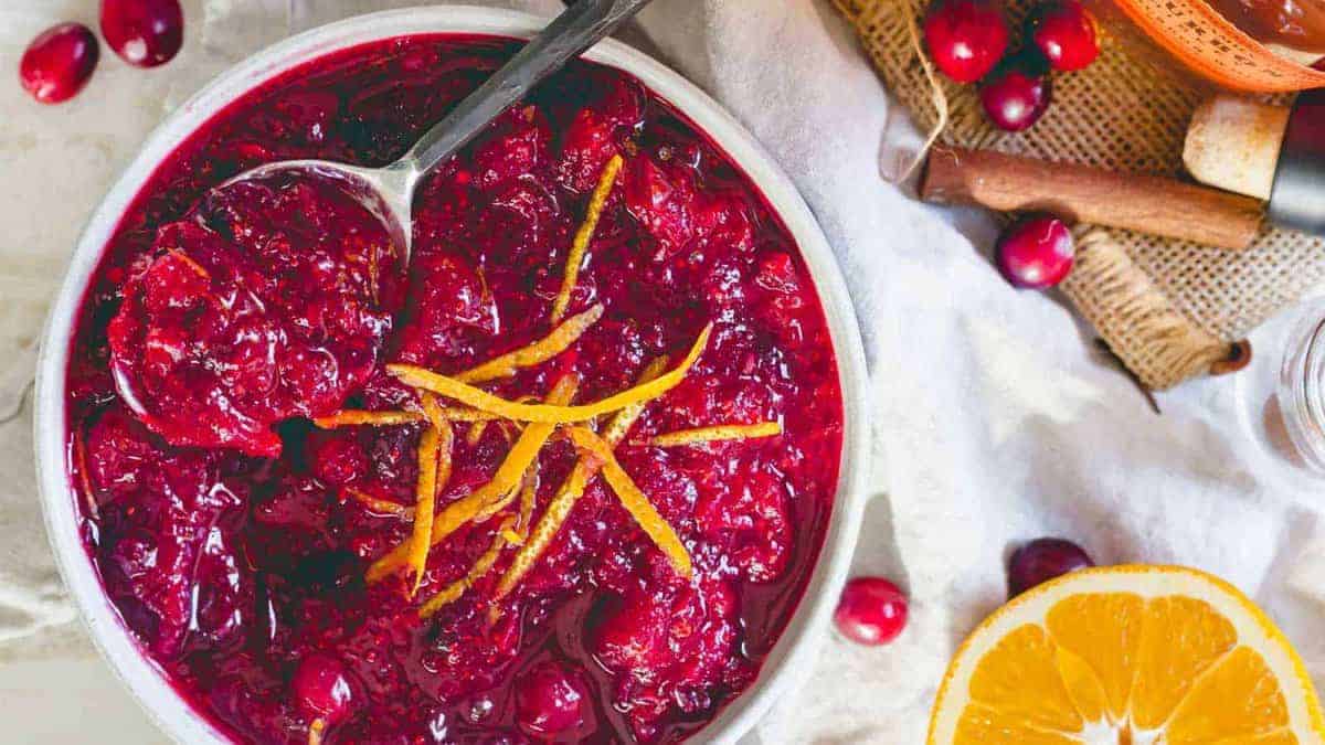 A bowl of cranberry sauce garnished with orange zest, with a spoon inside. Fresh cranberries and an orange half are on the side.