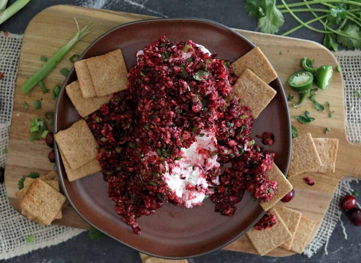 A plate of cream cheese topped with a chunky cranberry salsa, surrounded by square crackers on a wooden board with fresh herbs nearby.