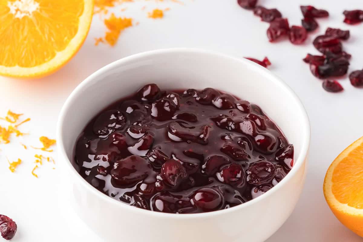 A white bowl filled with cranberry sauce sits on a white surface, surrounded by orange halves, zest, and a few dried cranberries.