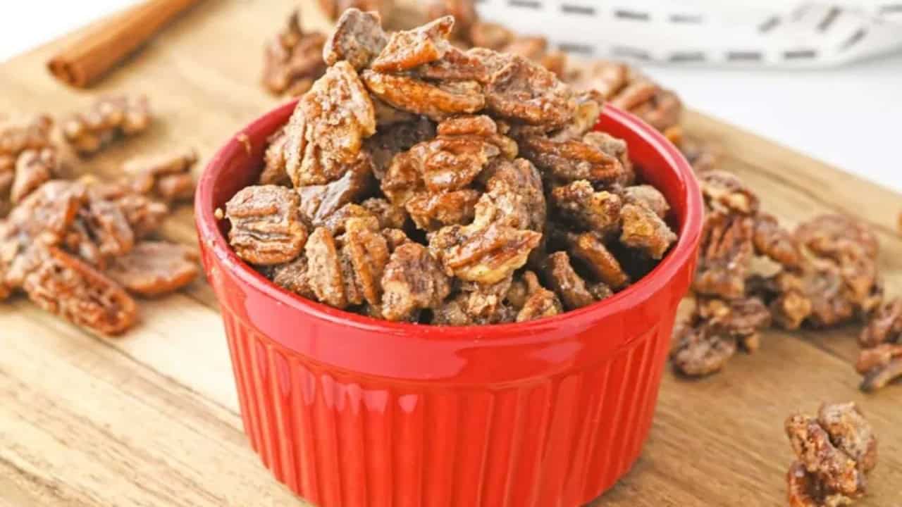 A red bowl filled with sugared pecans sits on a wooden surface, with more sugared pecans scattered nearby.