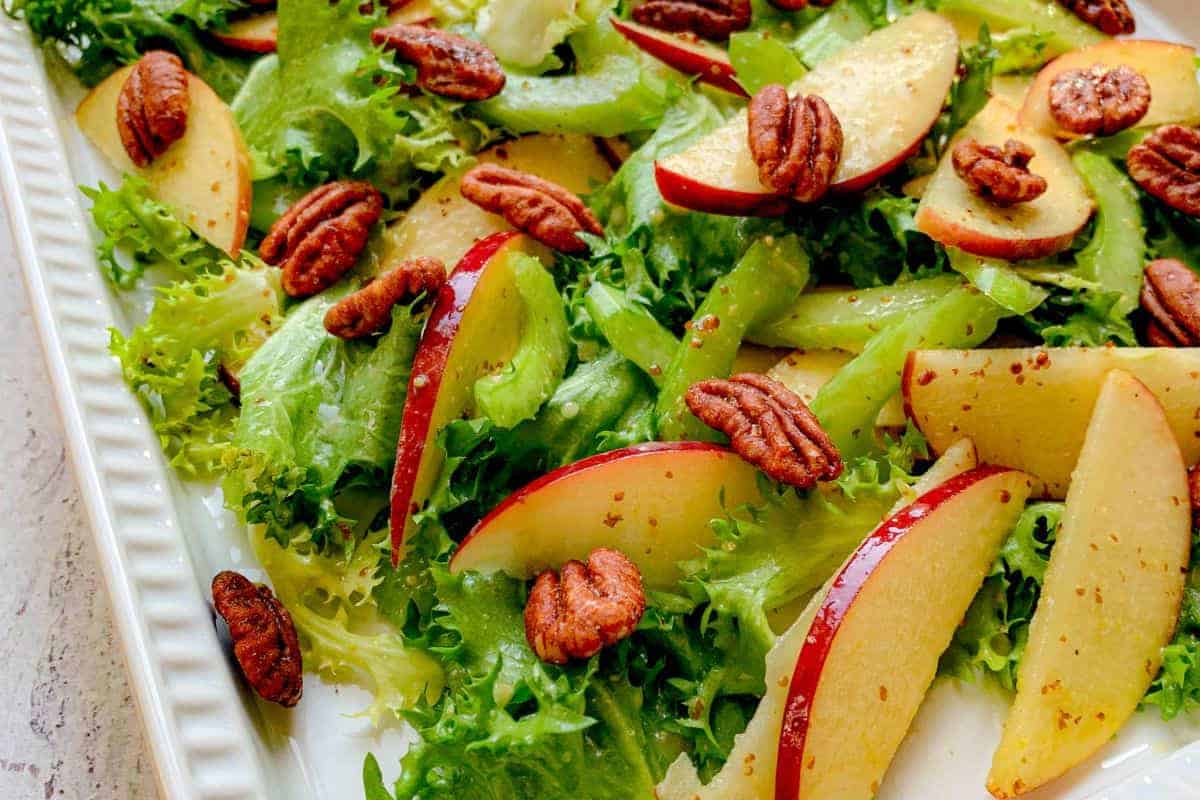A rectangular plate of green leafy salad topped with sliced red apples and whole pecans.