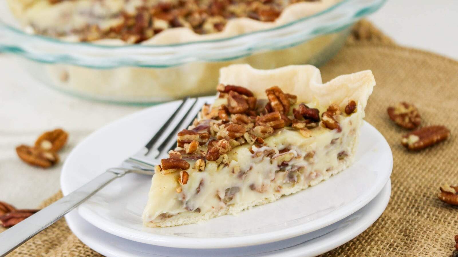 A slice of pecan cream pie on a white plate with a fork, with the remaining pie in a glass dish in the background.