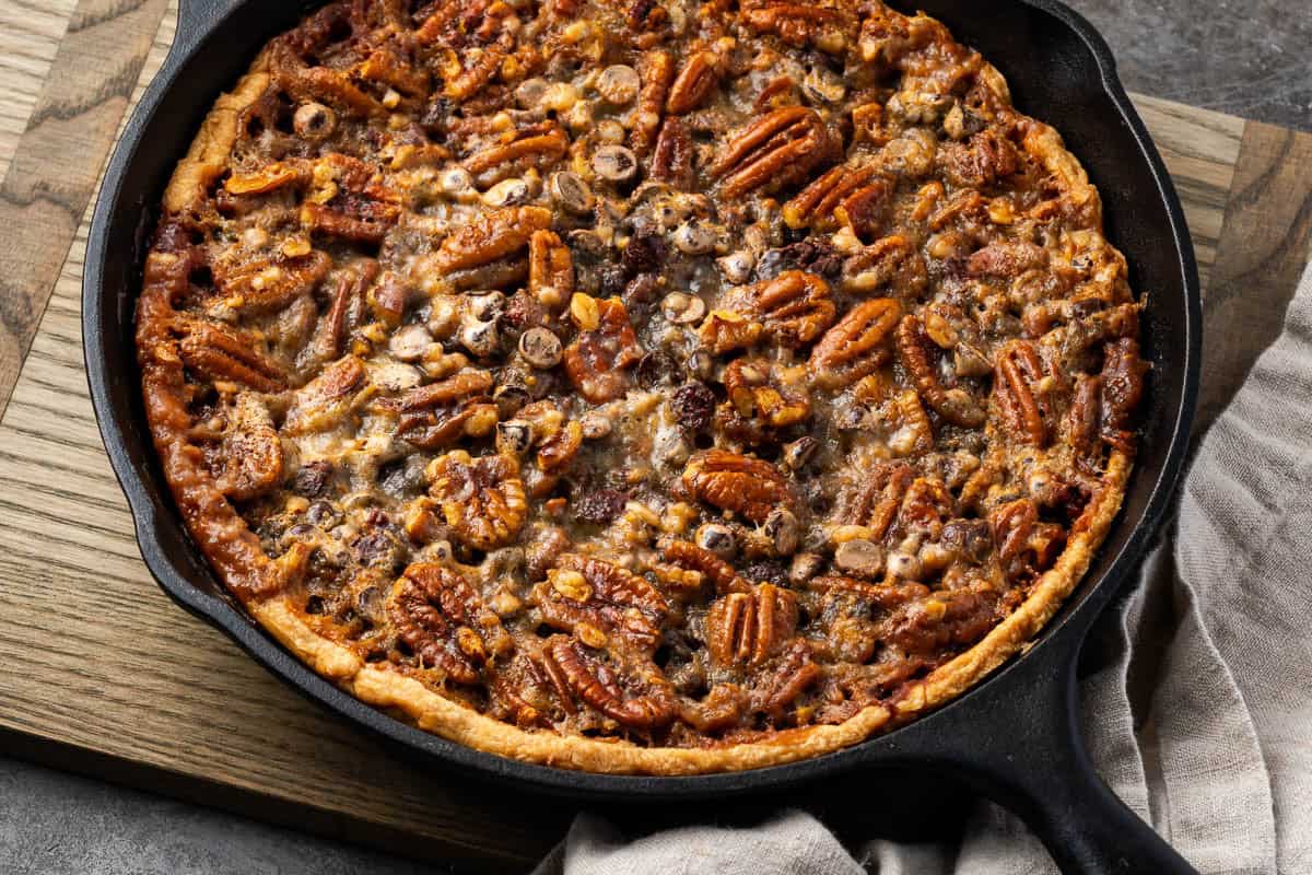 A baked pecan pie with a golden crust sits in a cast iron skillet on a wooden board, topped with whole pecans and nuts.