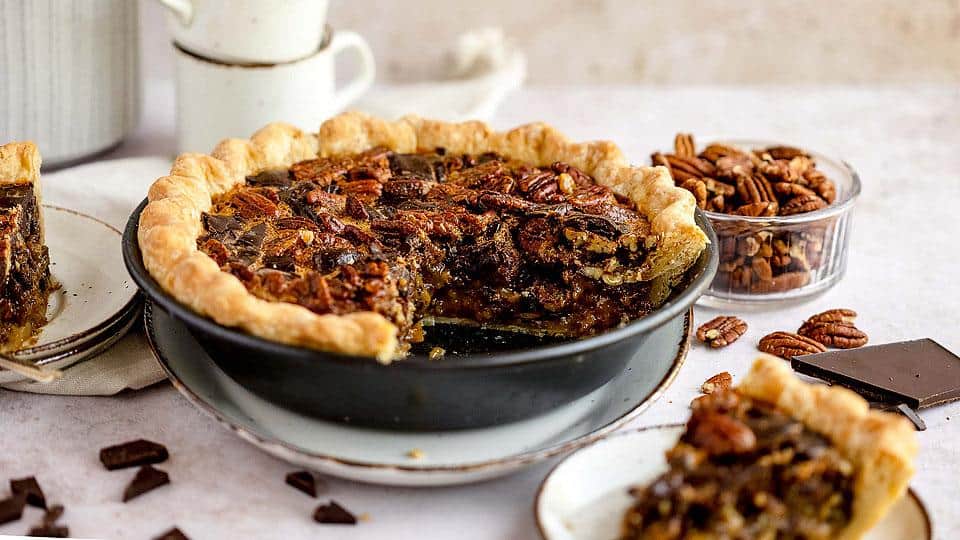 A pecan pie with a slice removed sits on a table, surrounded by pecans, chocolate pieces, and a small stack of cups.