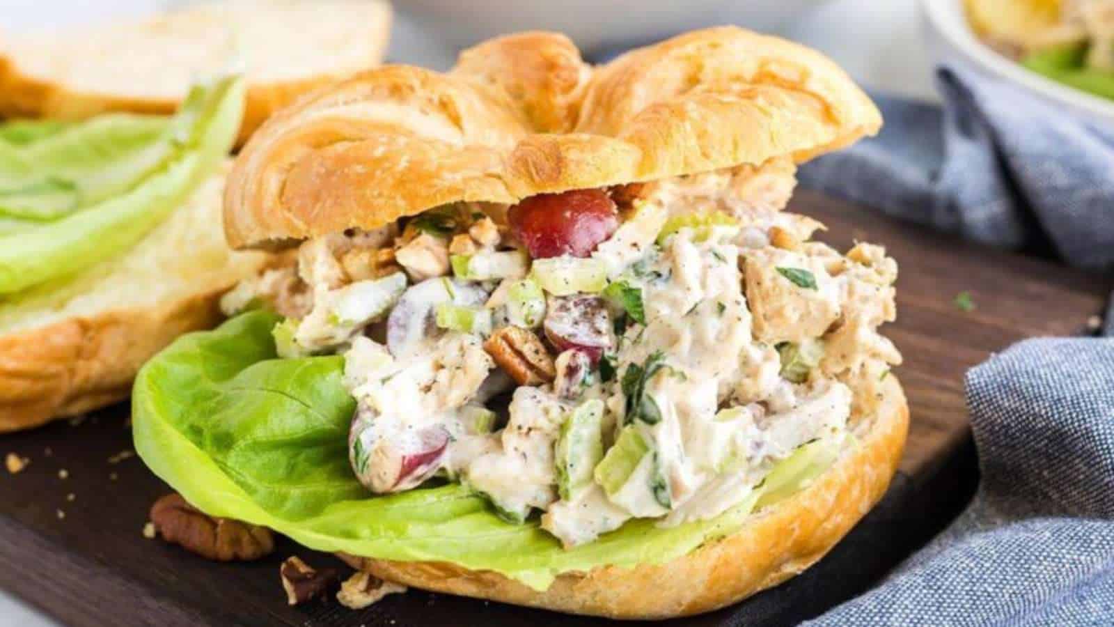 A croissant sandwich filled with chicken salad, grapes, celery, chopped nuts, and a lettuce leaf sits on a wooden board.