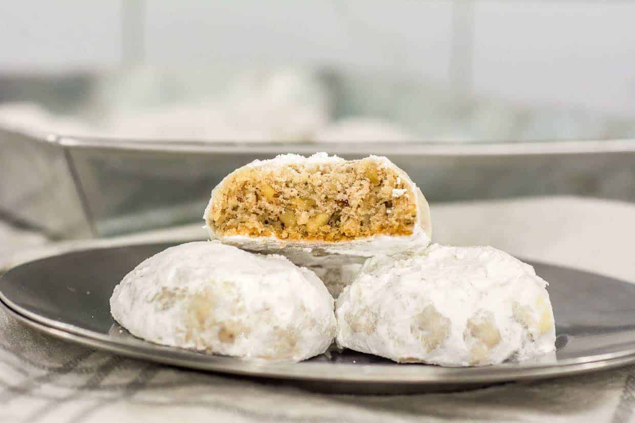 Three powdered sugar-covered cookies on a plate, with one cookie cut in half to show a crumbly, nut-filled interior.