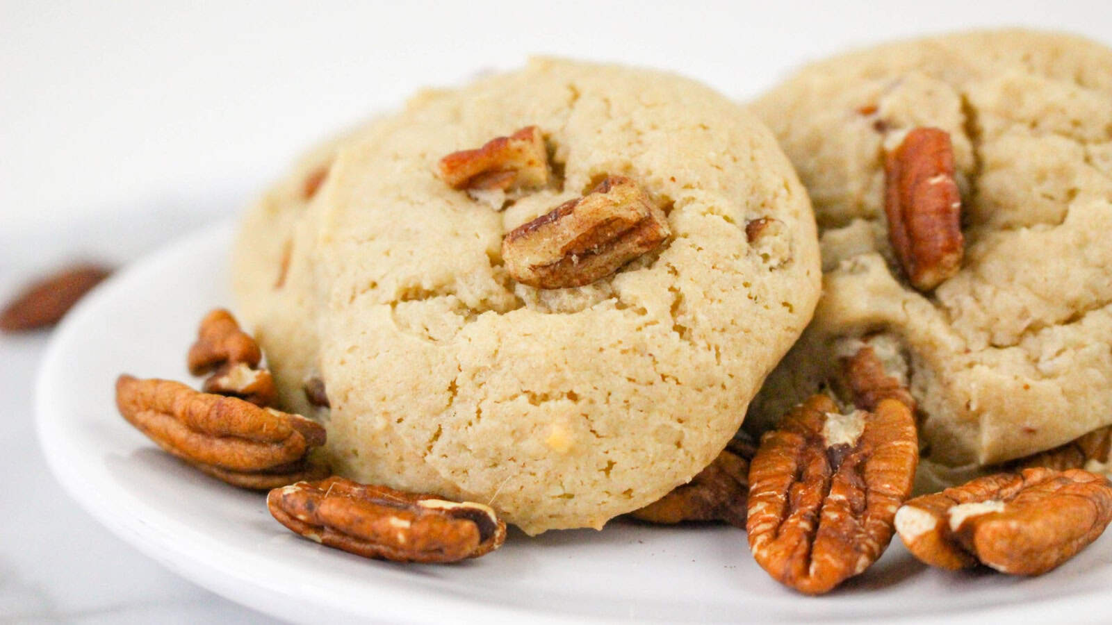 A close-up of pecan cookies on a white plate, with whole pecans scattered around them.