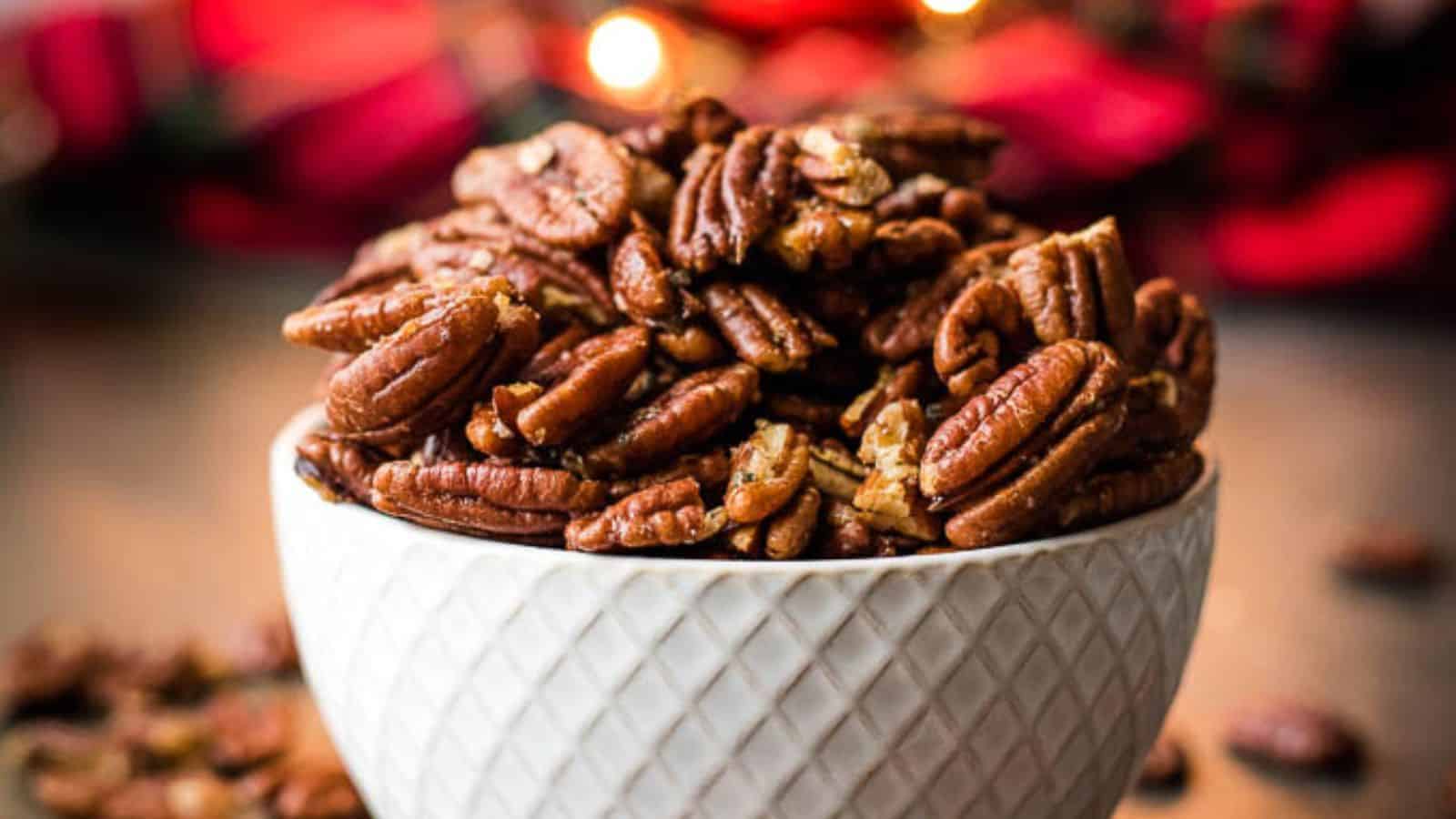 A white textured bowl filled with roasted pecans sits on a wooden surface, with a blurred background featuring red and orange lights.