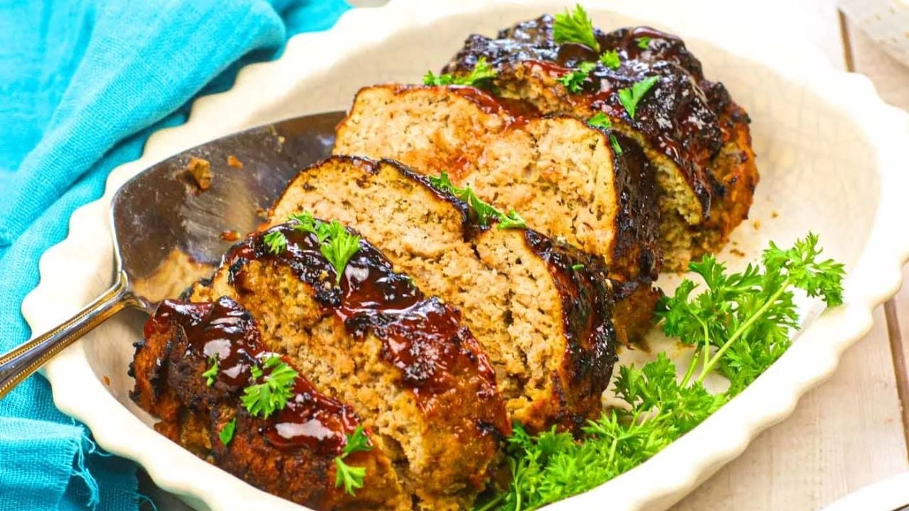 Sliced meatloaf topped with glaze, garnished with parsley, served in a white dish with a metal serving spoon.