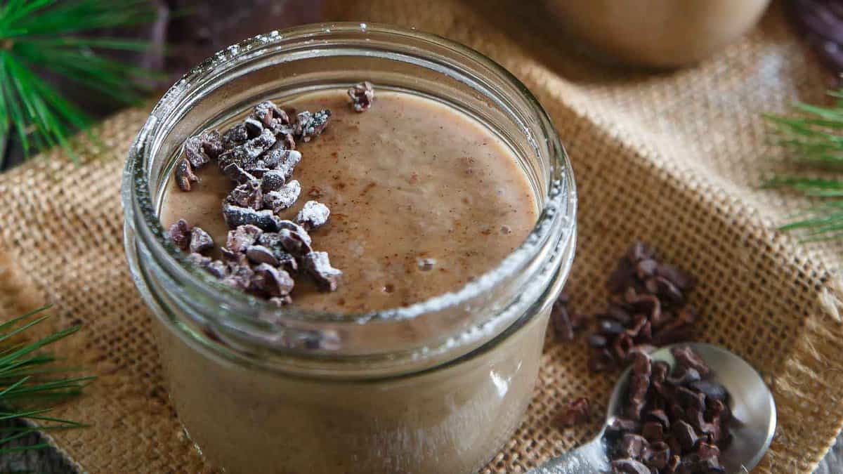A glass jar filled with chocolate mousse, topped with cacao nibs and powdered sugar, sits on a burlap surface next to a spoon and scattered cacao nibs.
