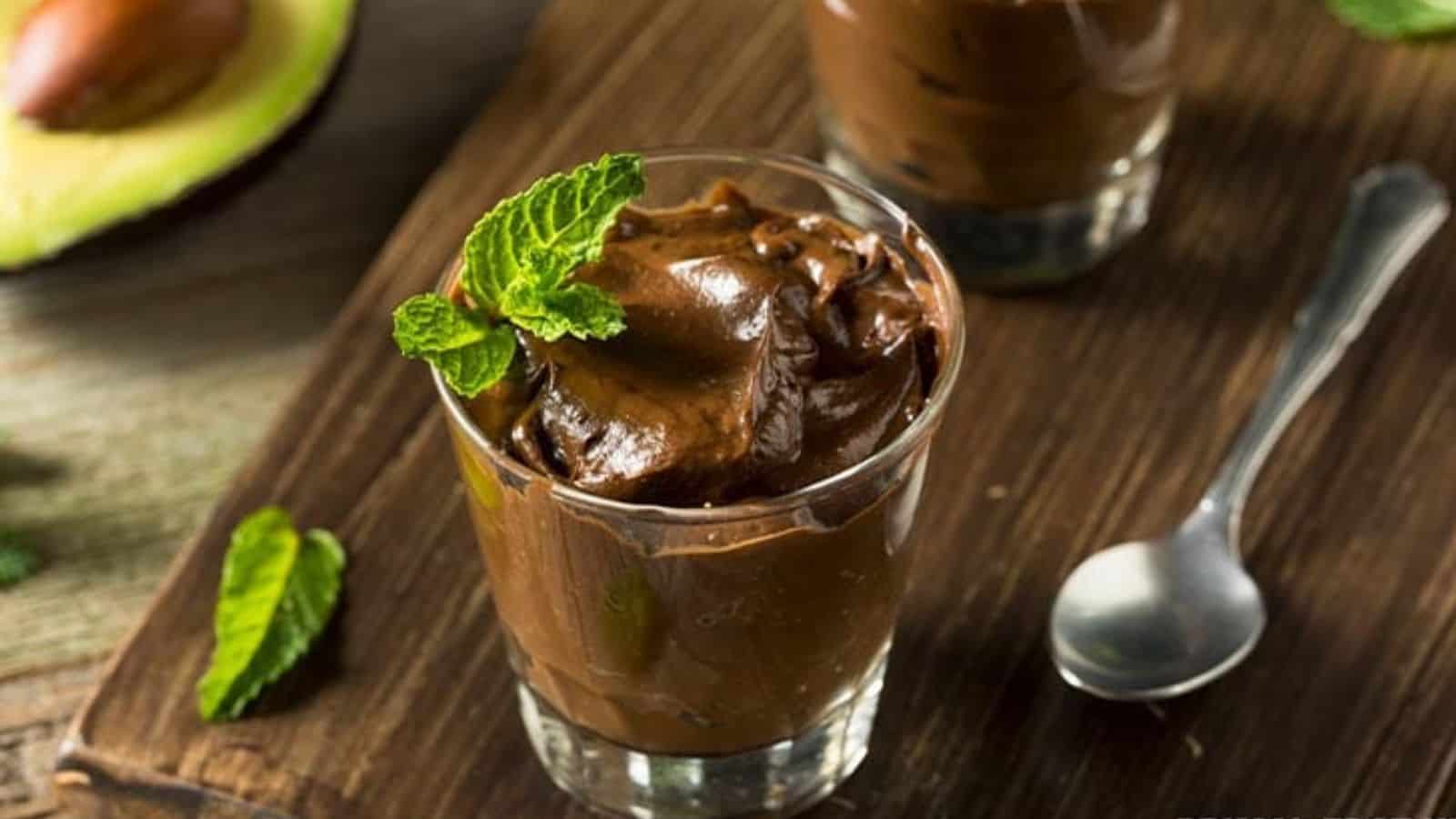 A glass of chocolate mousse garnished with a mint leaf sits on a wooden surface next to a spoon, with another glass and mint leaves in the background.