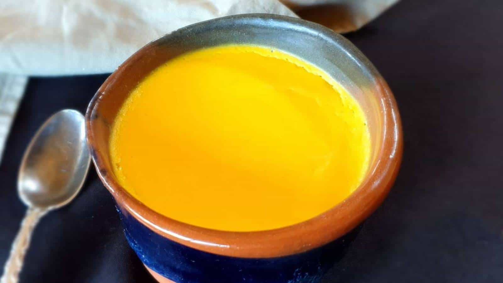 A clay cup filled with bright yellow turmeric milk sits next to a spoon on a dark surface.