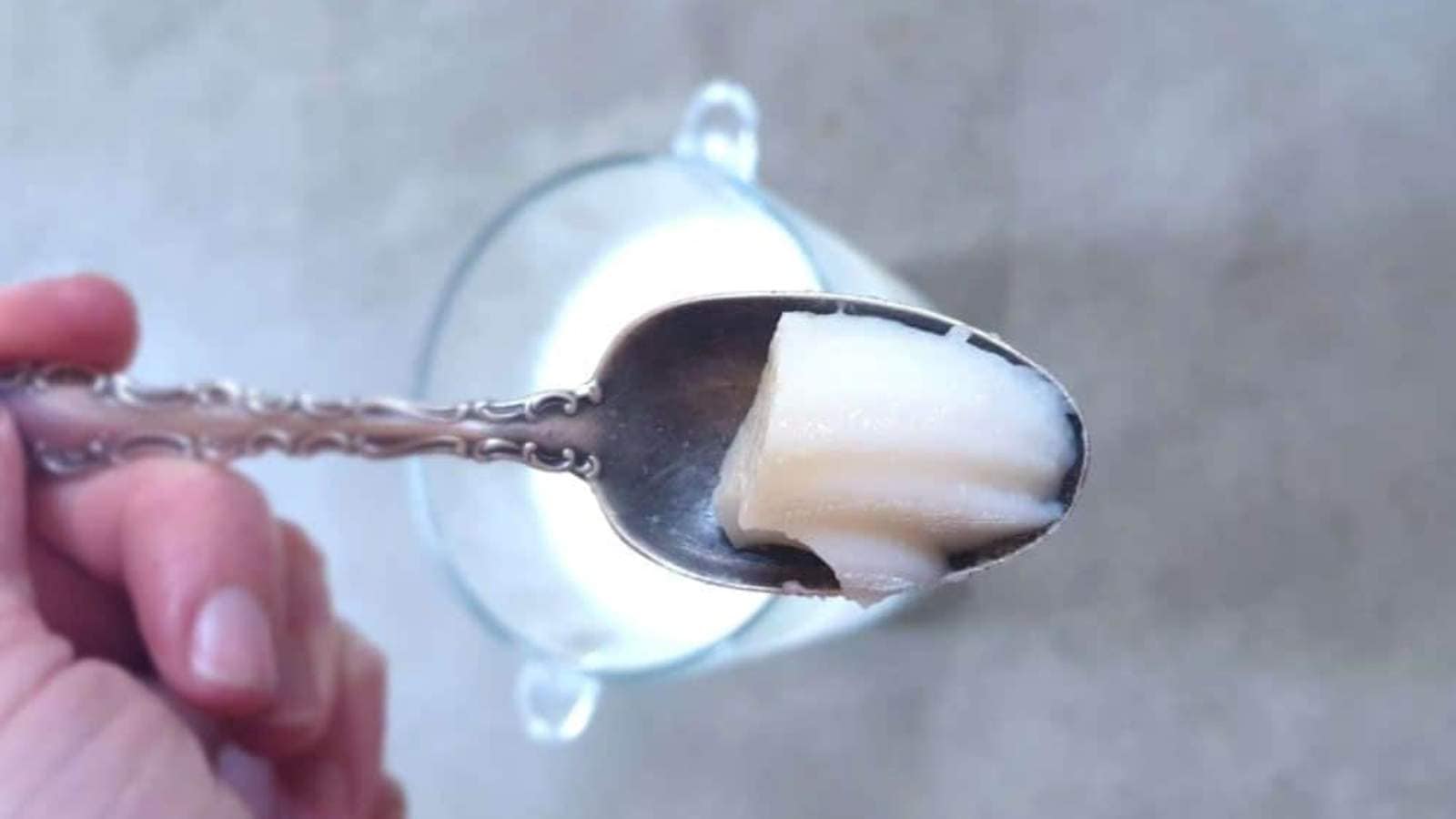 A hand holds a spoon of solid white substance above a glass cup filled with liquid on a gray surface.