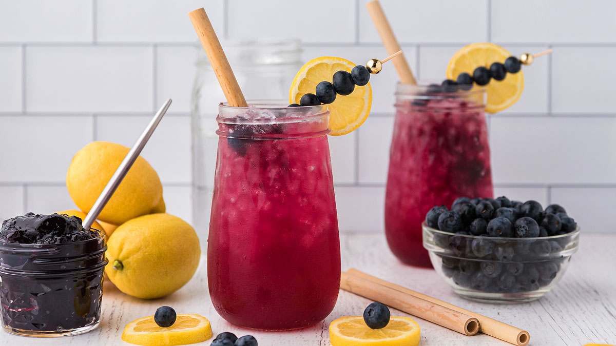 Two mason jar glasses of purple iced drink garnished with lemon slices and blueberries on skewers, next to fresh lemons, blueberries, jam, and bamboo straws on a white table.