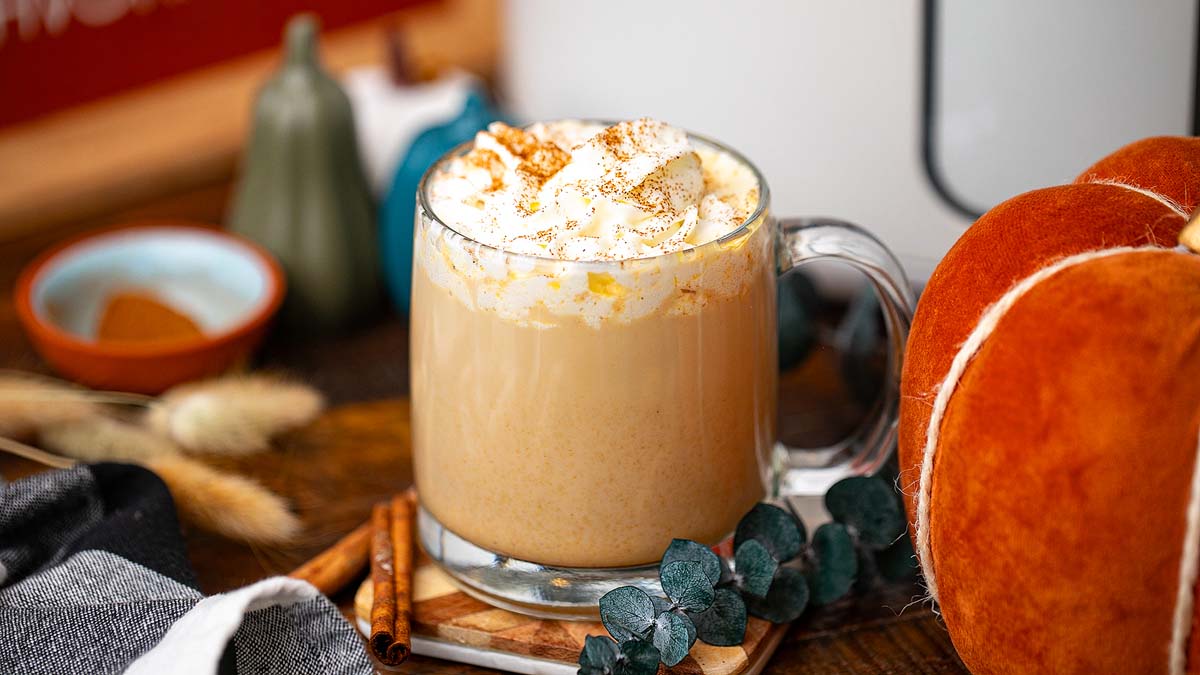 A glass mug filled with a creamy beverage topped with whipped cream and cinnamon sits on a wooden table, surrounded by pumpkins, cinnamon sticks, and decorative greenery.