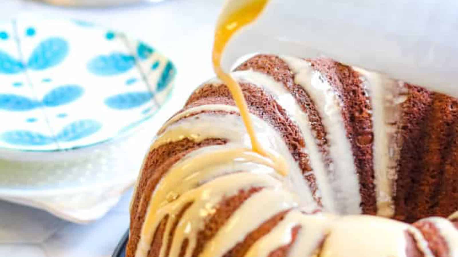 Close-up of icing being poured over a Bundt cake, with decorative plates visible in the background.