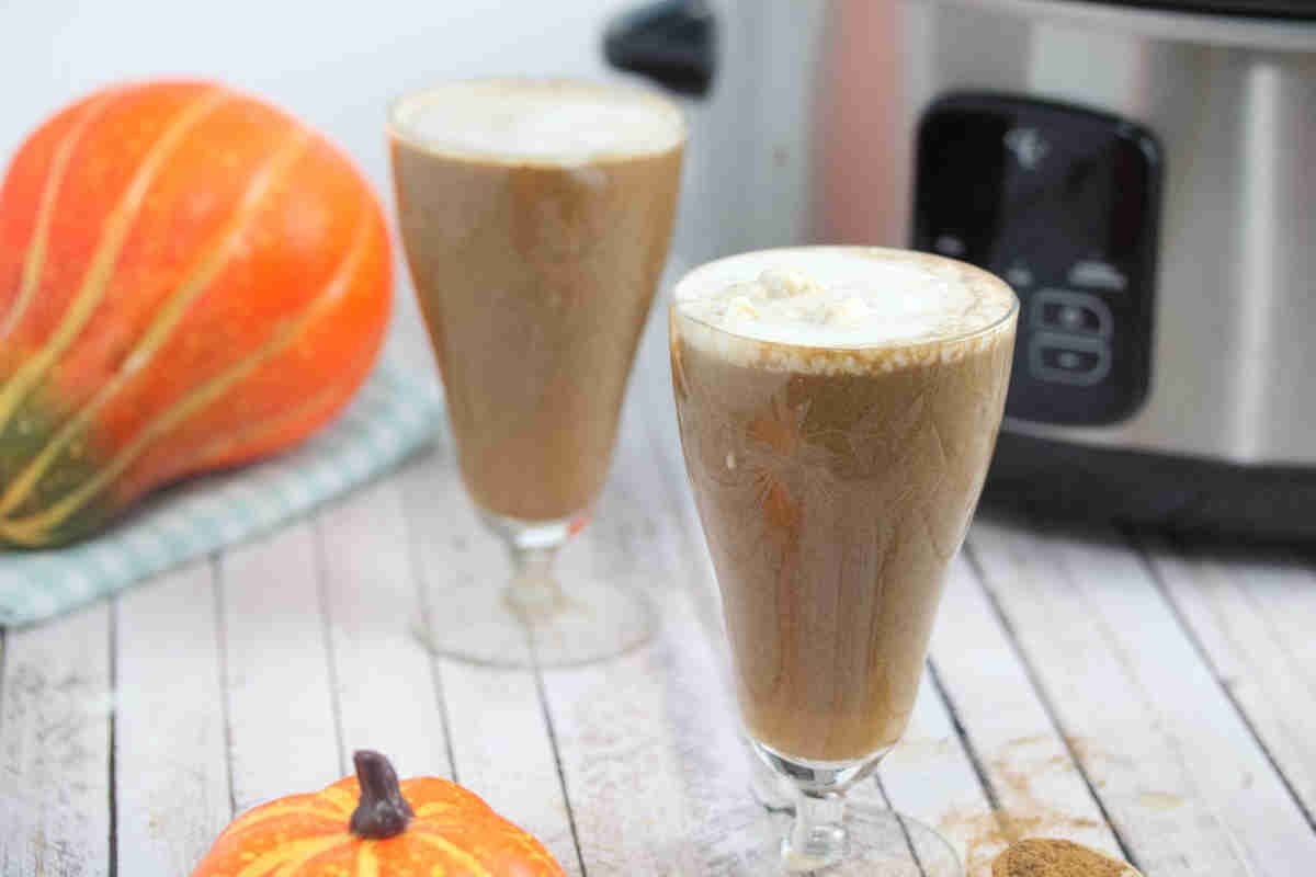 Two glasses of pumpkin spice latte with whipped cream on top sit on a white wooden surface next to a pumpkin and a slow cooker.