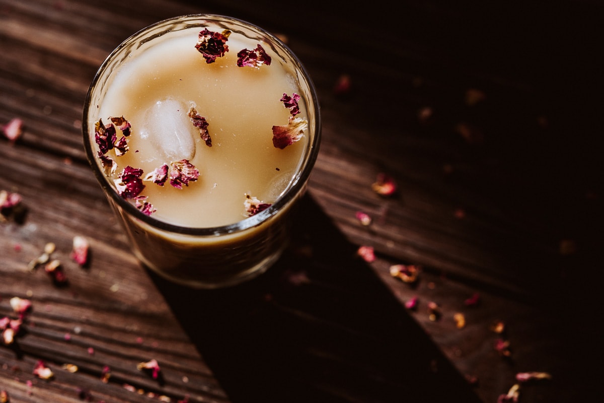 A glass of light brown iced beverage garnished with dried flower petals sits on a wooden surface with scattered petals around.