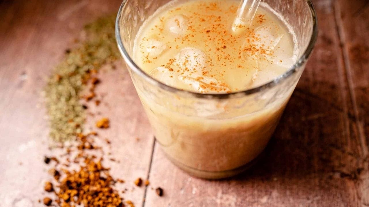 A glass of iced horchata with a straw, topped with ground cinnamon, sits on a wooden surface next to scattered spices.