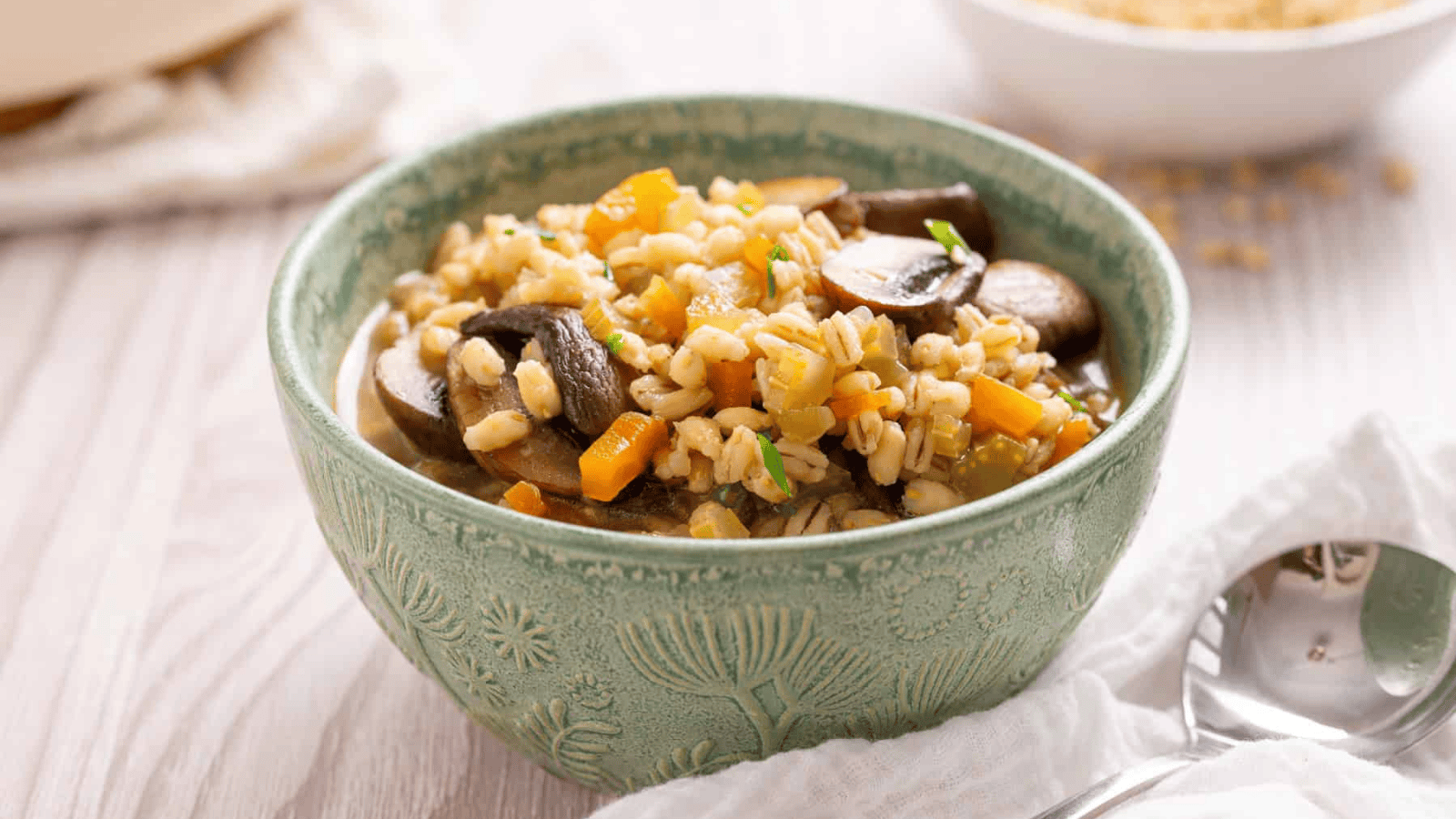 A green ceramic bowl filled with barley, mushrooms, and diced carrots sits on a white table next to a spoon and a white napkin.