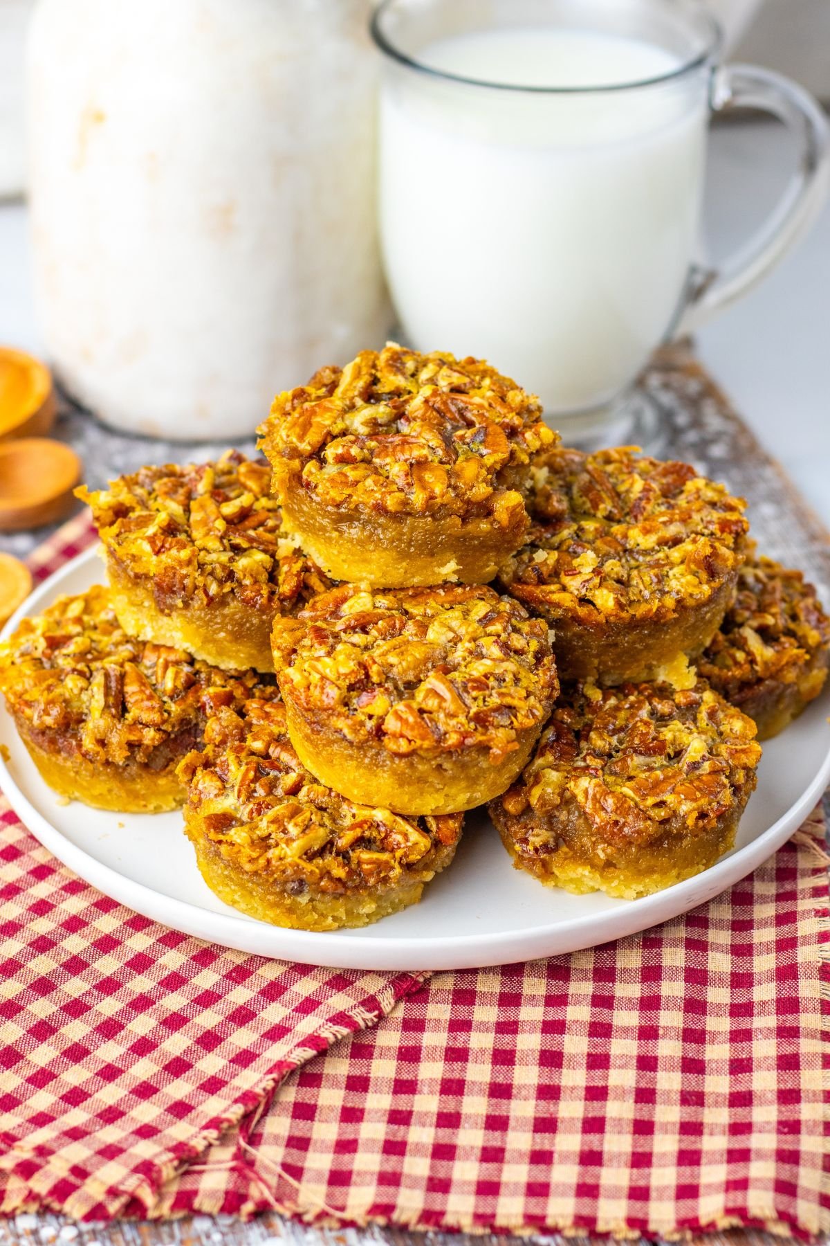 A plate of mini pecan pies stacked in a pyramid shape, with a glass of milk and a jar in the background, on a red checkered cloth.