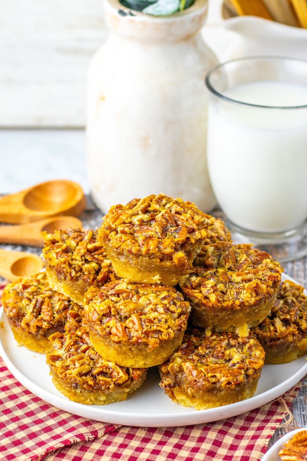 A plate of mini pecan pies is stacked on a table next to a glass of milk, a red checkered napkin, and wooden measuring spoons.