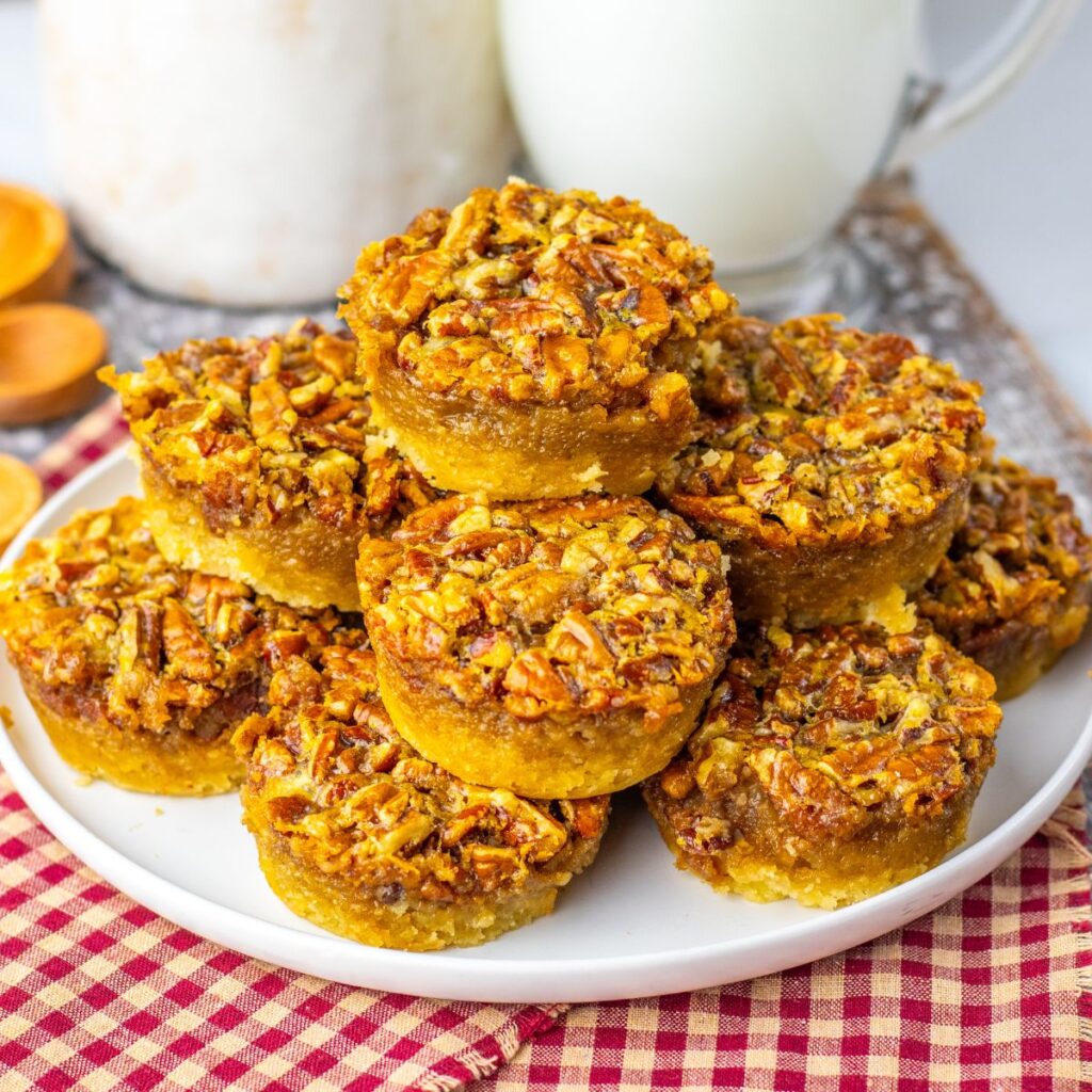 A plate of mini pecan pies stacked on top of each other, sitting on a red checkered cloth, with milk and a jar in the background.