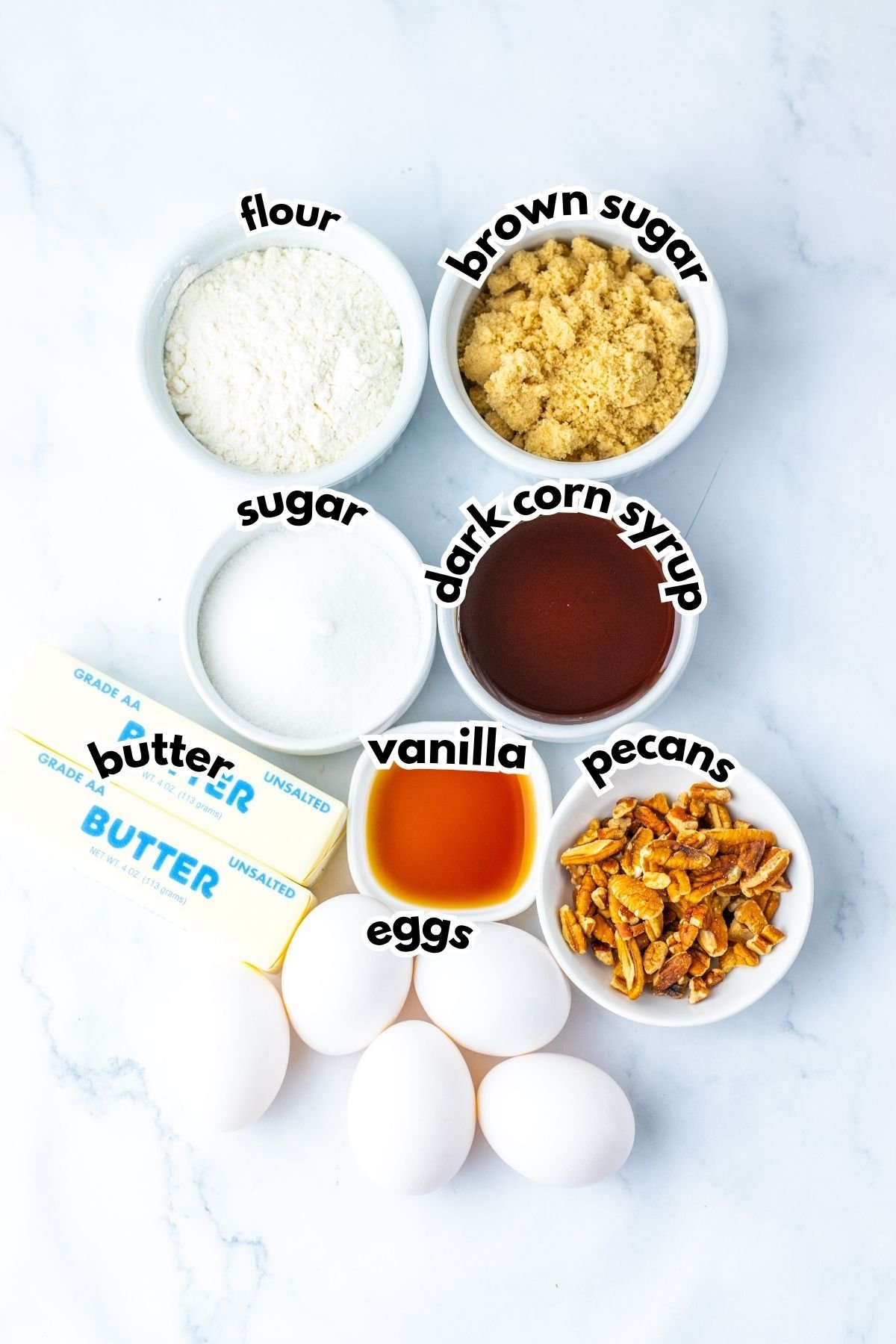 Ingredients for baking arranged on a white surface: flour, brown sugar, sugar, dark corn syrup, vanilla, pecans, unsalted butter, and four eggs.