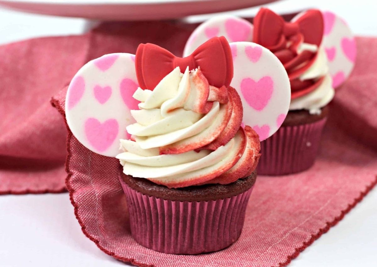 Two cupcakes with pink liners are topped with swirled white and pink frosting, heart-decorated fondant ears, and red fondant bows, sitting on a pink cloth.