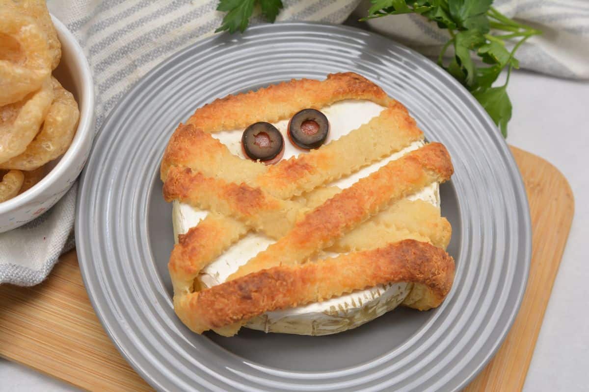 A round cheese wheel topped with pastry strips arranged like a mummy and two olive slices for eyes, served on a gray plate.