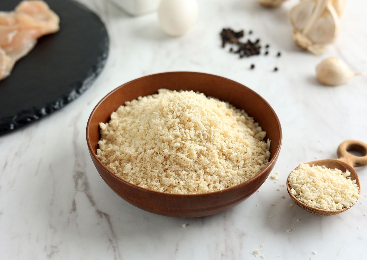 A brown bowl filled with panko breadcrumbs sits on a white surface, with a small wooden bowl of breadcrumbs and various cooking ingredients in the background.