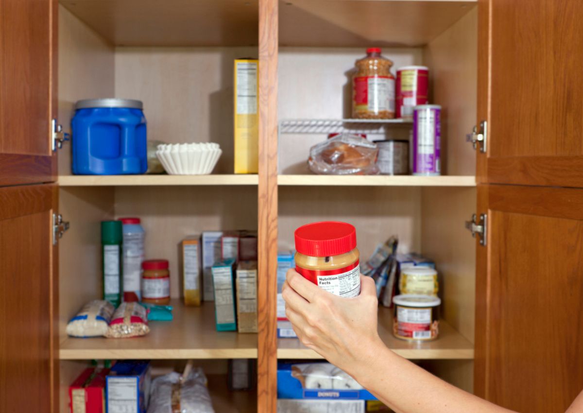 A hand holds a jar of peanut butter in front of an open pantry containing various food items, including cereal boxes, pasta, jars, and coffee filters.